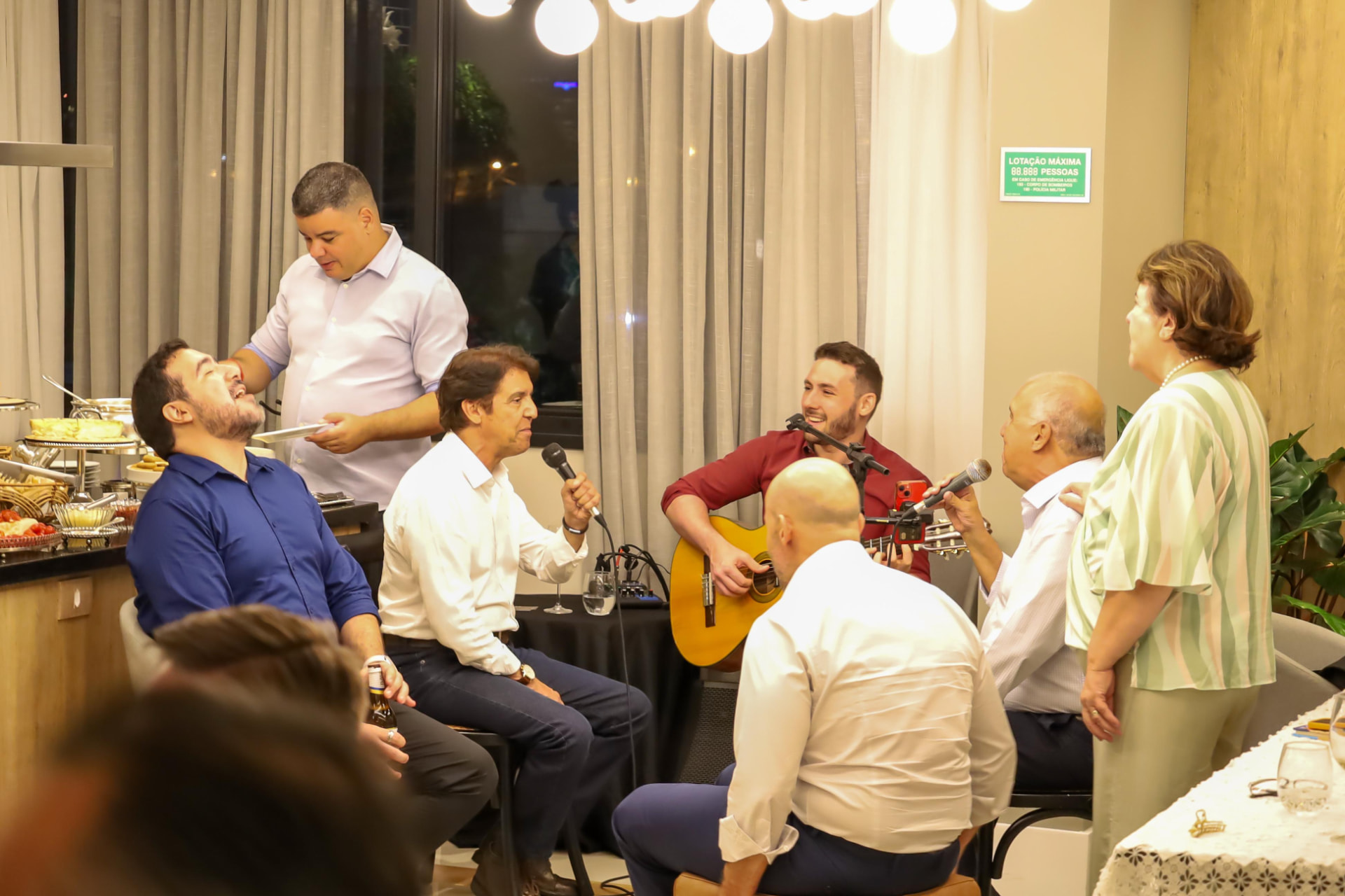 Roger Loeblein tocando violão e cantando em um evento corporativo, cercado de convidados